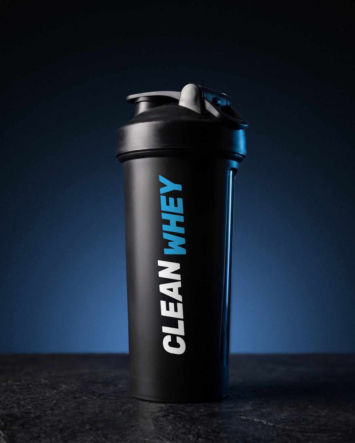 A matte black shaker bottle with a matching black lid, featuring the text 'CLEAN WHEY' printed vertically on the side in white and bright blue. The bottle is set against a dark blue gradient background on a dark, textured surface.